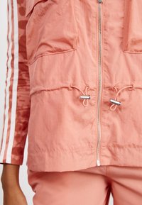 Person som bär en rosa jacka med framdragkedja, snoddar och vita ränder nerför ärmarna, i kombination med matchande rosa byxor.