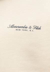 Λογότυπο Abercrombie & Fitch κεντημένο με γκρι κλωστή σε υφασμάτινο φόντο φωτεινού μπεζ με υφή, με τη φράση «Νέα Υόρκη, Ν.Υ.» κάτω σε κεφαλαία γράμματα.