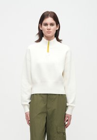 Osoba oblečená v bílém svetr s krátkým zipem a jemným logem, spolu s olivově zelenými cargo kalhotami, stojící na jednobarevném bílém pozadí.