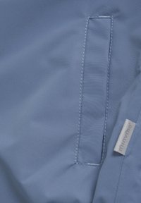 Tissu imperméable bleu clair avec une texture lisse, comportant une poche rectangulaire et une petite étiquette de marque cousue sur le bord.