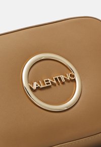 Borsa a tracolla in pelle tan con un logo circolare dorato con la scritta "VALENTINO" in rilievo, texture liscia e linee pulite.