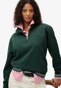 Groene rits-hoodie met geribbelde manchetten, met een verhoogd logo. Gedragen over een lichtroze overhemd met kraag en witte strepen.