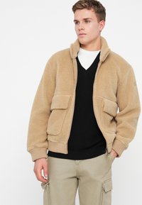 Casaco de fleece ceroso com fecho frontal, dois bolsos laterais e punhos canelados. Usado sobre um suéter preto com decote em V, combinado com calças cargo claras.