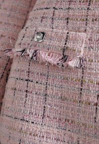 Rosa tweed-stoff med et geometrisk mønster, som har metalltråder og en sølvfarget firkantet knapp. Slitte kanter med dekorative detaljer.