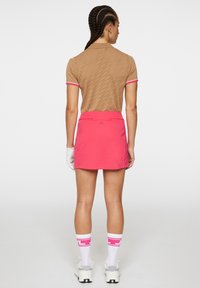 Bruine polo met patroon en roze bies, gecombineerd met een roze skort. Witte sokken met roze accenten en witte sportschoenen maken de outfit af.