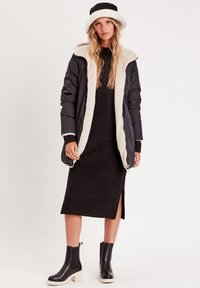 Abrigo largo negro acolchado con forro crema y capucha; vestido negro de canalé con abertura lateral; botas negras hasta los tobillos con suela crema. Diseño sencillo.