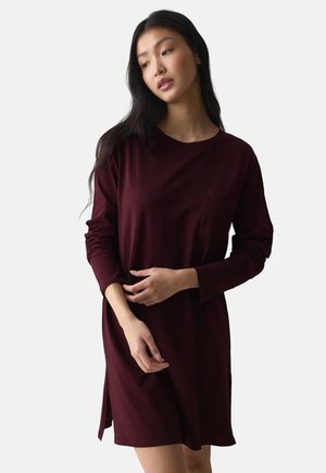 Mujer con pelo largo y negro que lleva un vestido de manga larga de color burdeos oscuro, de pie con las manos suavemente entrelazadas, mirando ligeramente hacia abajo y hacia un lado.