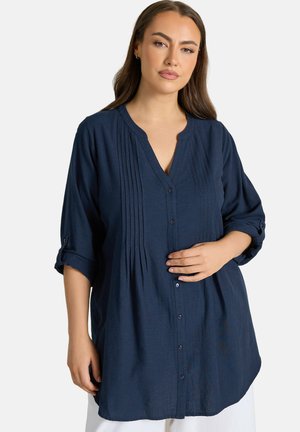 Vrouw met lang bruin haar, gekleed in een marineblauwe tuniek met knopen en opgerolde mouwen, die met één hand op haar buik rust, witte achtergrond.