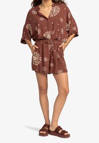 Salopette marron à motifs avec des manches courtes, boutonnage sur le devant, taille élastique avec cordon de serrage, et poches. Portée avec des sandales à plateforme marron.