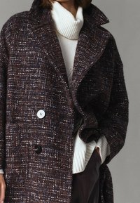 Uldblandingsfrakke i mørkebrun med hvide striber, med klapkrave, store knapper og løs pasform, lagdelt over en hvid turtleneck.