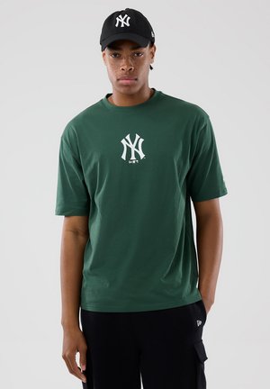 Joven con camiseta verde oscuro de los New York Yankees y gorra negra, de pie frente a un fondo gris claro liso.