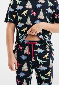 Pyjama set met zwarte stof met kleurrijke patronen van dinosaurussen en kerstbomen. T-shirt met korte mouwen en broek met drawstring in rood accent.