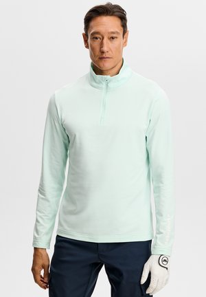 Mand iført lyseblå kvart-lyn pullover og mørke bukser, holder en hvid golfhandske i højre hånd og står foran en ensfarvet baggrund.