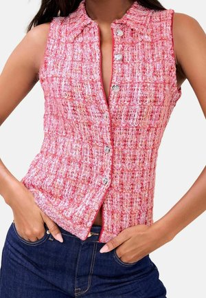 Mouwloze roze getextureerde knoopvest gedragen met donkerblauwe spijkerbroek, handen rustend in de voorzakken.