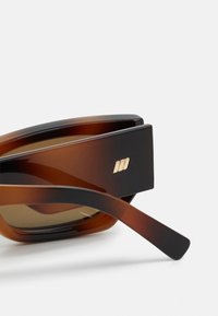 Sunglasses med överdimensionerade brun gradientbågar, svarta detaljer och en gyllene logotyp på sidan. Texturerad yta med böjda skalmar.