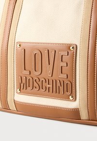 Τσάντα χειρός από καμβά σε χρώμα ταμπά και εκρού με υφασμένη επιφάνεια. Διαθέτει εμφανές καφέ δερμάτινο λογότυπο με ανάγλυφη την επιγραφή "LOVE MOSCHINO". Λεπτομέρειες με χρυσά καρφιά και ραφές.