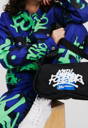 Giacca con motivo graffiti blu e verde con bottoni a pressione, borsa a tracolla nera con logo bianco e sneakers bianche con accenti blu.