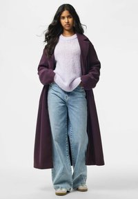 Jeune femme aux longs cheveux foncés porte un manteau violet, un pull violet clair, un jean large bleu et des baskets blanches sur un fond uni.