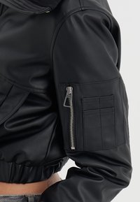Bomber jacket de cuero negro con cuello de canalé, bolsillo con cremallera en la manga y cinturilla elástica. Textura suave y diseño ajustado.