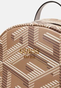 Hnědý syntetický batoh s geometrickým vzorem v krémové a tmavších odstínech. Zlaté logo "GUESS EST 1981" na přední straně. Texturovaný povrch.