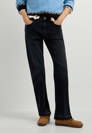 Persoon draagt zwarte rechte jeans, blauwe trui, bruine mocassins, staand met handen in de zakken tegen een effen lichte achtergrond.
