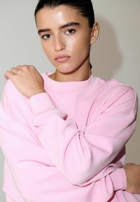 Svetloružová mikina s voľným strihom a rebrovanými manžetami, so zaobleným výstrihom a bezšvovým dizajnom ramien. Hladká textúra.