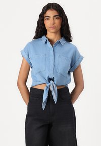 Camisa cropped em chambray azul claro com mangas curtas dobradas, frente com botões e detalhe de nó na cintura. Dois bolsos no peito.