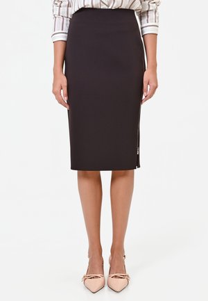 Femme portant une jupe crayon noire taille haute avec une fermeture éclair latérale, associée à une chemise rayée et des escarpins pointus couleur nude.