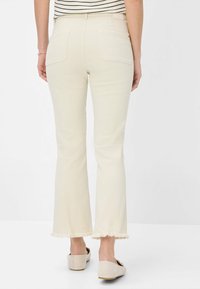 Light beige flared jeans featuring a raw fringe hem and back pockets, made of denim fabric. The model wears neutral-colored shoes.