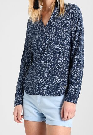 Vrouw draagt een navy blouse met lange mouwen met wit bladmotief en lichtblauwe shorts met opgerolde zoom, staand tegen een effen achtergrond.