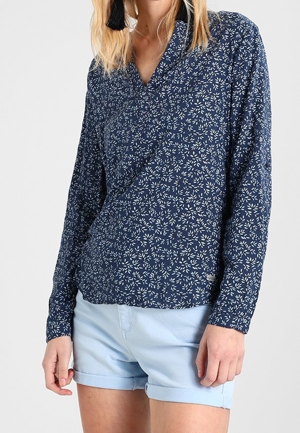 Femme portant un chemisier bleu marine à manches longues avec un motif de feuilles blanches et un short bleu clair à ourlet roulotté, debout devant un fond uni.