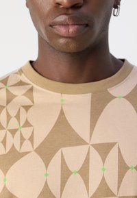 Beige T-Shirt mit kurzen Ärmeln, das ein geometrisches Muster in Braun und Grün zeigt, mit Rundhalsausschnitt und weichem Baumwollstoff.