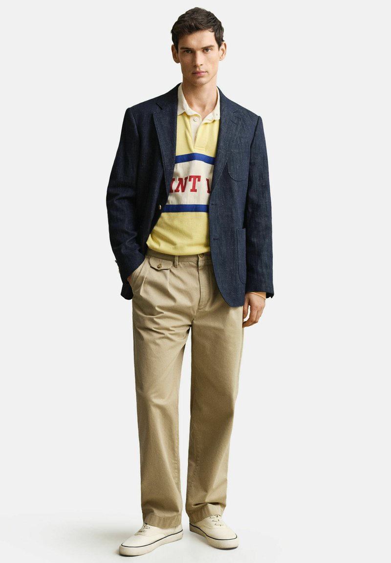 Junger Mann trägt dunkelblauen Blazer, gelbes Poloshirt mit roten und blauen Streifen, beige Hose und weiße Sneakers, steht mit den Händen in den Taschen.