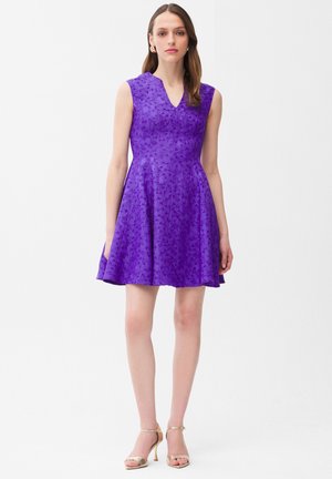 Kvinde iført en ærmeløs lilla kjole med blomsterprint, V-udskæring, skørt med vidde og gyldne højhælede sandaler, stående mod en hvid baggrund.