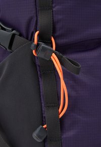 Gros plan d'un sac à dos violet, avec des bretelles noires, un cordon orange et une boucle. Tissu texturé avec des sections en maille et lisses visibles.