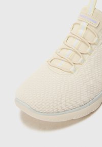 Beige athletic shoe with a textured mesh upper, elastic laces, and a rubber sole. Features subtle white and yellow accent lines.