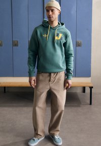 Grön hoodie med gula dragsnören och logotyp, kombinerad med tan färgade löst sittande byxor och mintgröna sneakers. En enkel beanie framhäver stilen.