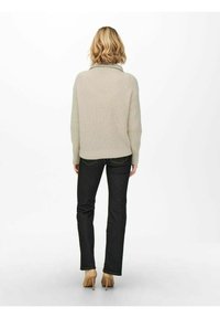 Pull en tricot beige clair avec une coupe décontractée, associé à un jean foncé et des talons beiges, vu de dos sur un fond uni.