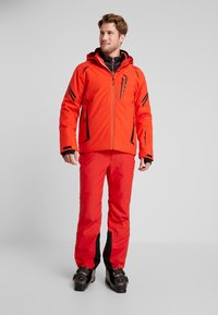 Ljust orange skidjacka med svarta accenter och flera fickor med dragkedja, tillsammans med röda byxor med svarta nedre paneler och skidskor.