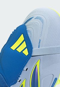 Gros plan d'une chaussure de football bleue et jaune néon avec une tige texturée, des lacets élastiques, et les inscriptions "NONOSTRIKE" et "ARTIFICIALGRASS".