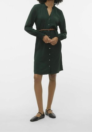 Femme portant une robe boutonnée vert foncé avec des poignets froncés, une ceinture tressée marron et des ballerines Mary Jane noires, debout devant un fond uni.