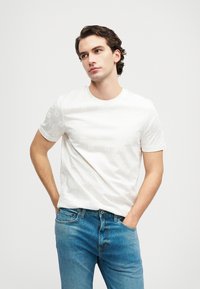 Cămașă albă din bumbac cu un model floral texturat, guler rotund, mâneci scurte, combinată cu blugi din denim bleumarin. Modelul stă cu mâinile în buzunare.