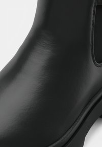 Black, glossy ankle boot with smooth, shiny surface, side cutout, and rounded toe. Features contrasting matte sole details.