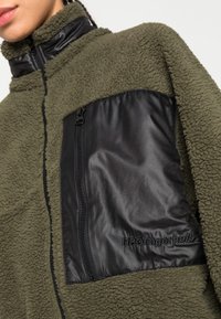Giacca in pile verde con una tasca in nylon nera lucida con zip, colletto alto e un leggero motivo testurizzato sul tessuto.