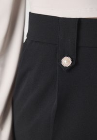 Pantalon noir taille haute en tissu lisse, accentué d'un grand bouton en perle, avec une taille ajustée et un devant plat.