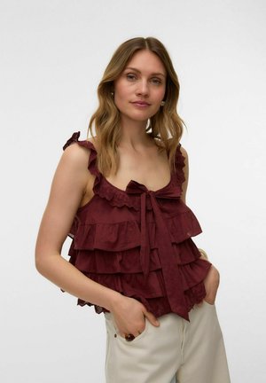 Femme aux cheveux ondulés porte un haut bordeaux sans manches à volants avec un nœud, mains dans les poches d’un pantalon crème, expression neutre, fond blanc.