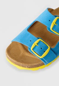 Μπλε δερμάτινο σανδάλι με δύο κίτρινες αγκράφες, καμπυλωτό καφέ πέλμα και κίτρινη άκρη στη σόλα, φωτογραφημένο σε λευκό φόντο.