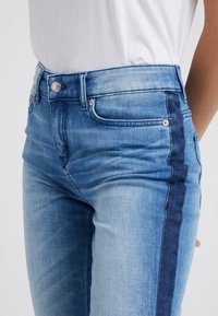 Vaqueros de tiro alto de color azul con un patrón desvanecido, con una franja de azul más oscuro a lo largo del costado, fabricados en denim, con bolsillos frontales y un botón plateado.