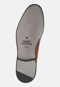 La suola della scarpa in pelle marrone presenta una soletta grigia con marchio impresso. Punta arrotondata con tacco in gomma nera e borchie dorate per una presa migliore.