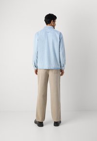 Light blue denim shirt with long sleeves, button cuffs, and stitched hem, worn over beige pants and black shoes. Plain background.